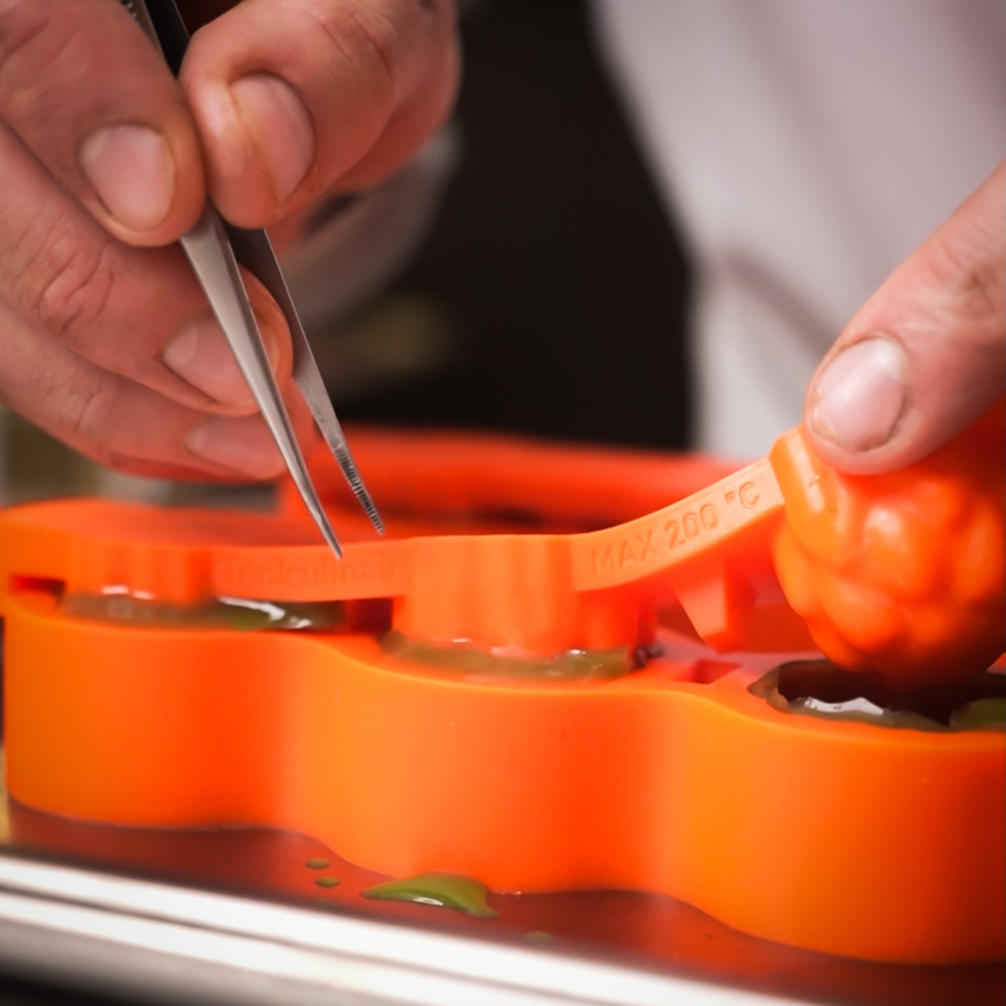 Close-up of mise en place showing tweezers removing an artichoke shape from a Toolculinair orange silicone mold, with the mold marked up to 200°C.