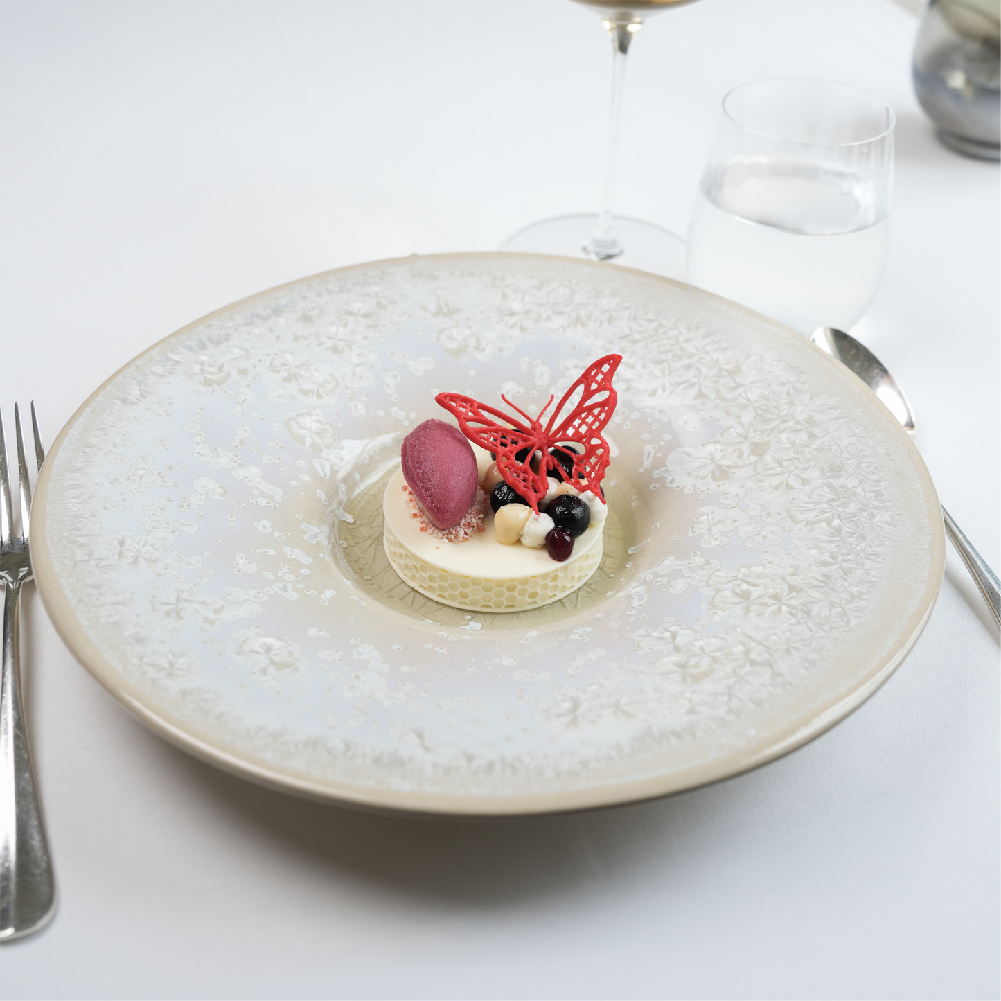 Atmospheric dessert presentation at The Moerbei by Michael Corpel, featuring a plated dessert topped with a red butterfly tuile, served with wine glass and cutlery in a fine dining table setting